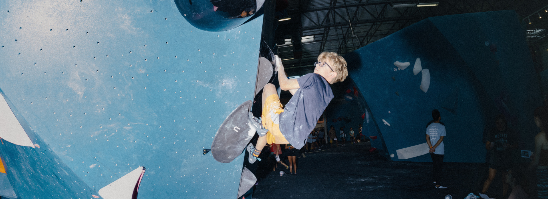 A person wearing a gray shirt, yellow shorts, and climbing shoes is bouldering indoors on a large blue climbing wall in Brooklyn, with Youth Teams and other climbing structures visible in the background.