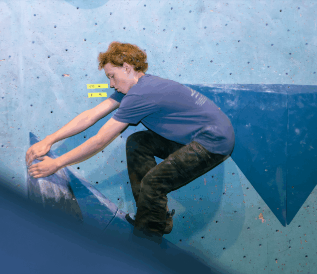 A person with short, curly hair climbs an indoor bouldering wall at an Austin gym, gripping large blue holds, wearing a blue t-shirt and dark pants, focused on the route labeled V4/5—perfect inspiration for local Youth Programs.