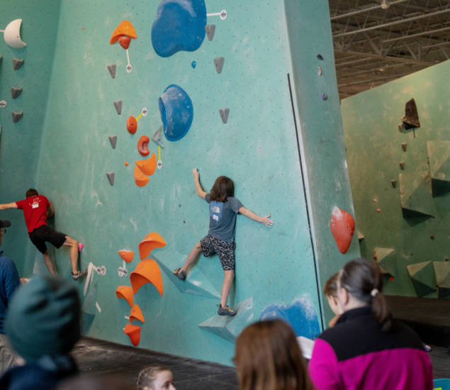 Children climb colorful holds on an indoor bouldering wall at a well-lit Minneapolis gym while adults watch from below. Blue and orange grips cover the high wall, perfect for kids training with the Climb Team.