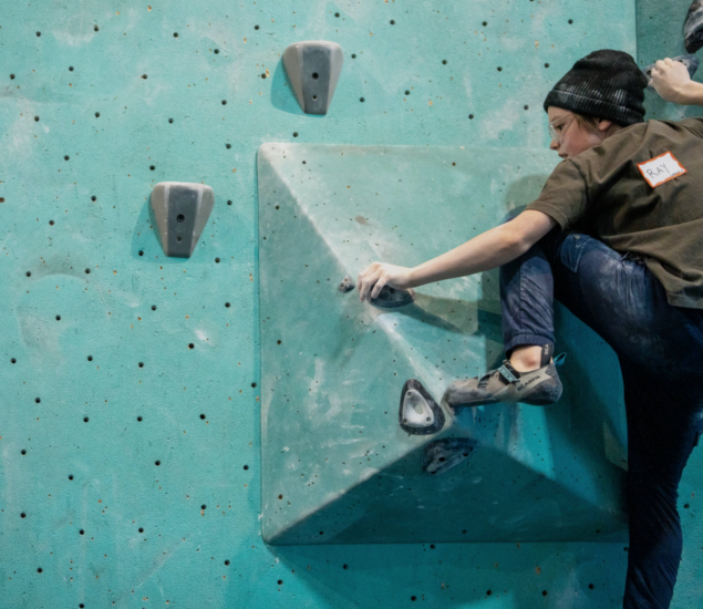 A person wearing a beanie and climbing shoes is scaling an indoor climbing wall in Minneapolis, gripping large geometric holds and using chalk on their hands for grip—perfect training for the Climb Team.